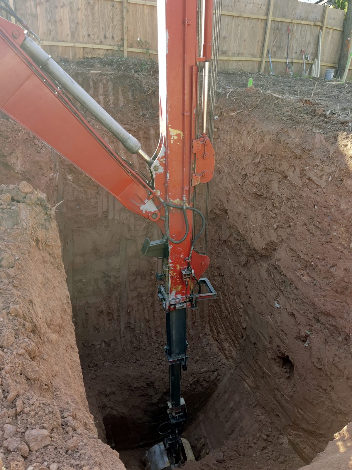 Telescoping long reach arm digging a deep, narrow pit beside a fence
